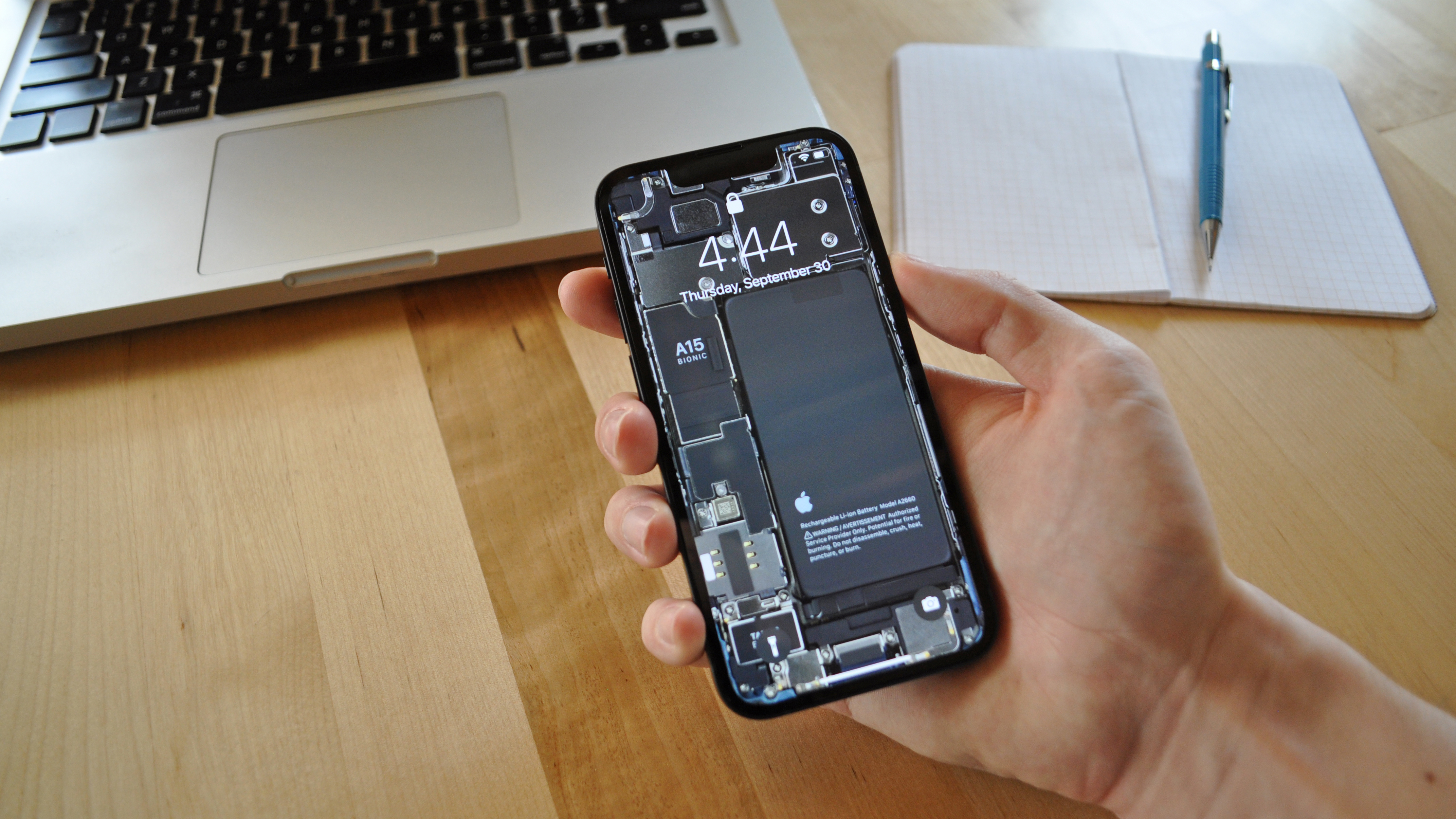 An iPhone 13 mini with internals wallpaper applied is held over a desk containing a MacBook and a notebook with pencil.