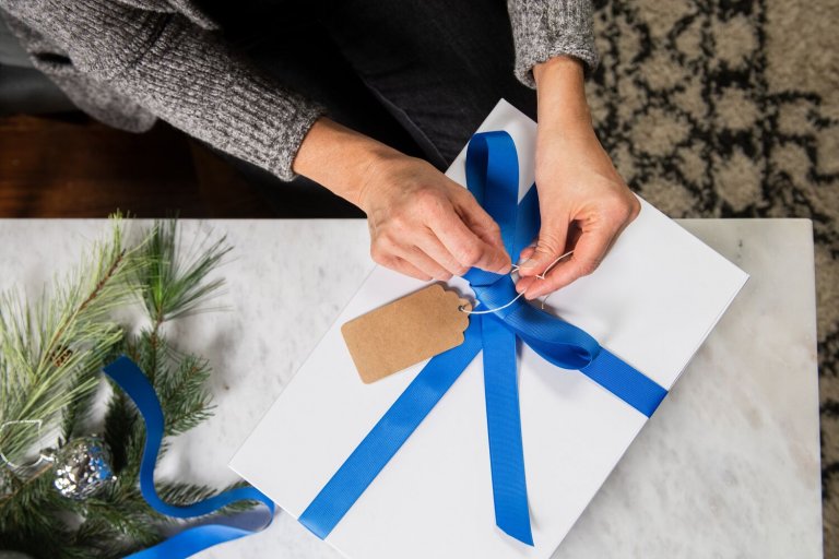 Hands tie a blue bow around a white box with a kraft paper tag. Evergreen branches are in the background.