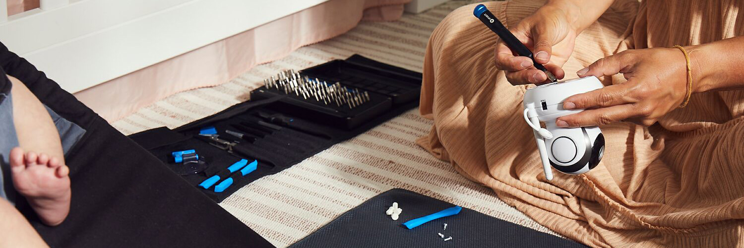 A person fixes a baby monitor with an iFixit Pro Tech Toolkit laying on the ground next to an infant in a baby bouncer.