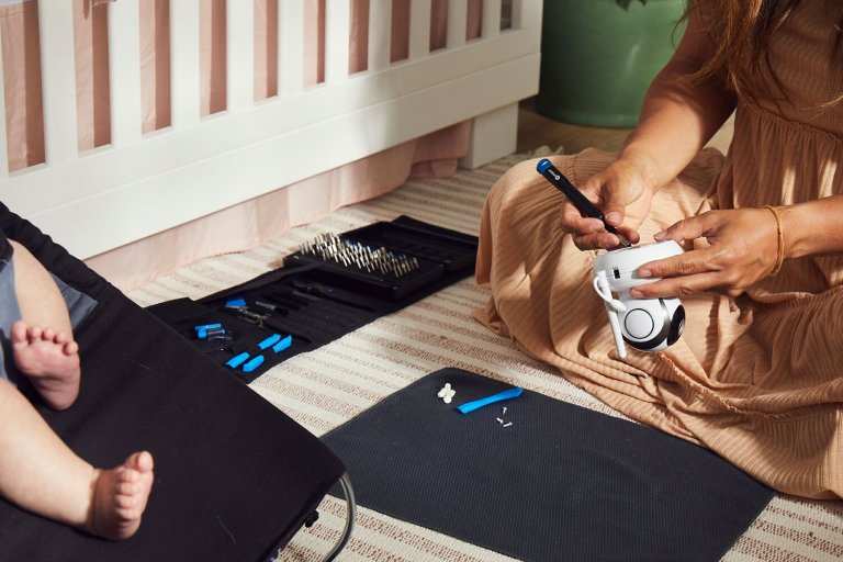 A person fixing a baby monitor while sitting on the ground next to an infant in a baby bouncer.