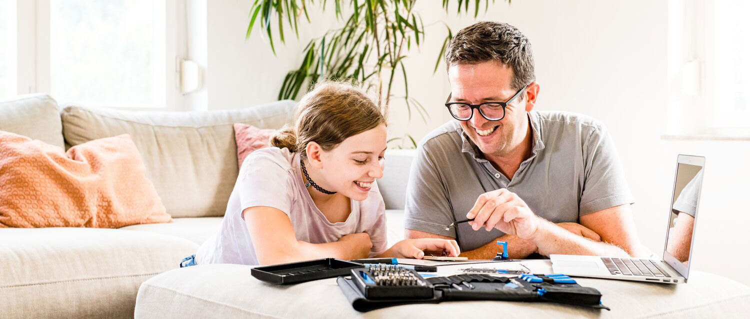 A dad and daughter smile while doing a repair in a brightly lit living room, with tools and a laptop in the foreground.