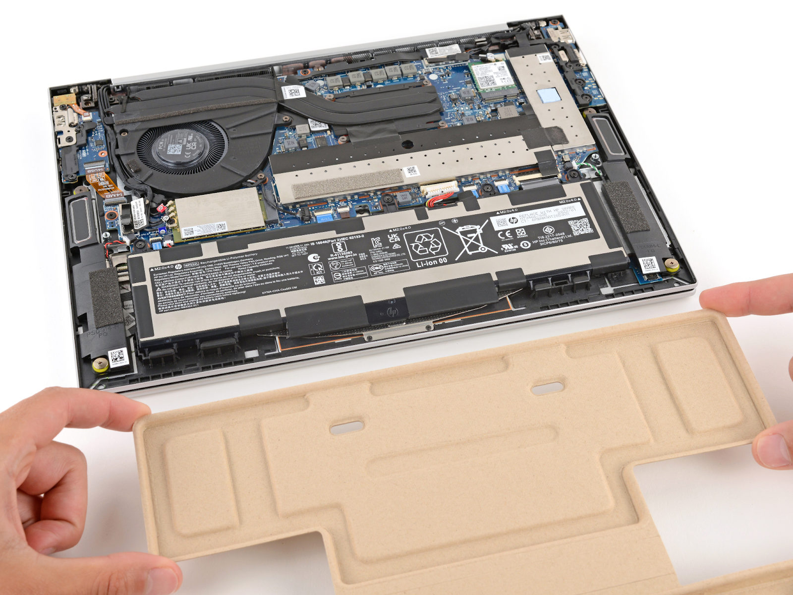Ein HP EliteBook G9 liegt kopfüber auf einer weißen Oberfläche, die untere Abdeckung ist entfernt. Zwei Hände halten das Revivekit aus Pappkarton bereit, um es auf dem Akku anzubringen. 
