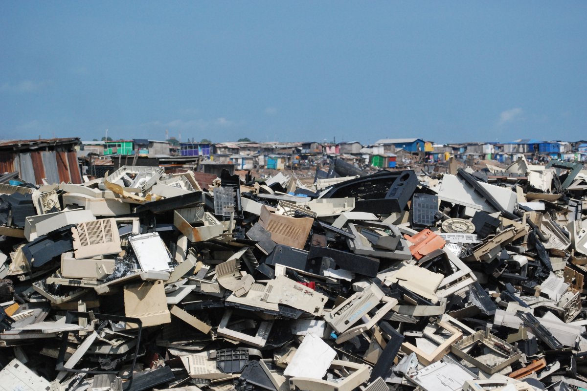 A photo of a mound of plastic and electronic waste with small, colorful buildings in the background and a blue sky overhead.