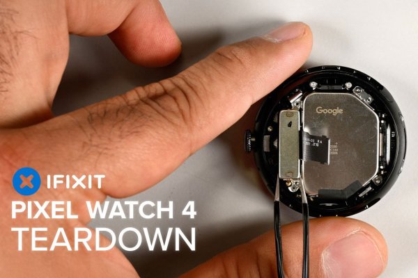 Close-up of an opened Pixel Watch 4 during teardown—one hand steadies the case as tweezers lift a small component beside a metal shield labeled “Google.”